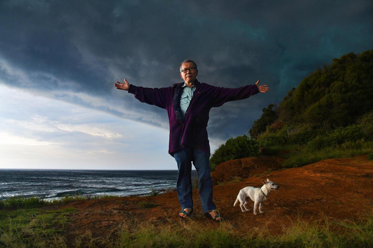 An Asian man, WIlliam Yang, at a coast setting. He has his arms spread out wide. There is a small white dog next to him.