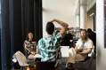 small scale performance: three people sitting around a table at a library. One person has their back to the camera and is posing to the other two who are the audience members in a small scale performance work.