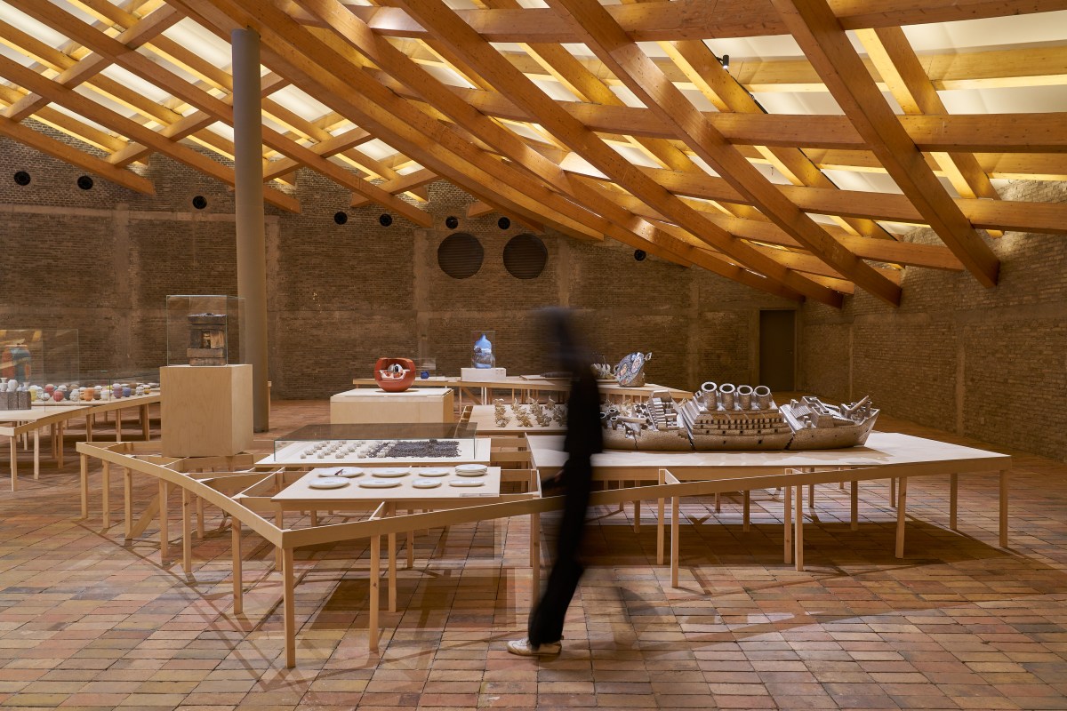 ‘The Ways of Clay: Select Award-Winning Works from the International Ceramics Festival Mino of the Museum of Modern Ceramic Art, Gifu, Japan’, installation view at UCCA Clay. An architectural space with earthy brick floor and slanted timber slits are roof, housing a dozen ceramic pieces. A black shadow of a figure is captured at the centre of the photo. 