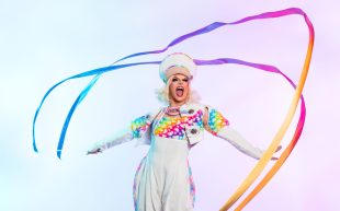 A performer in a white jumpsuit with rainbow trim. Swirling around them are two rainbow ribbons.