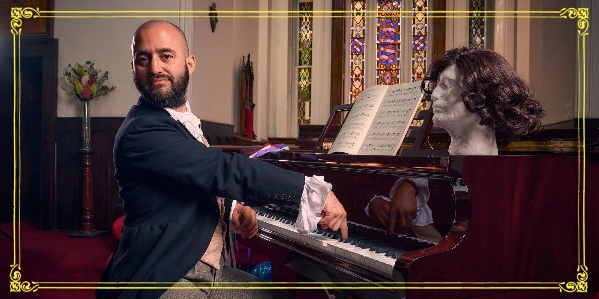 A bald man sitting at a piano. On top of the piano is a brown wig on a bust.