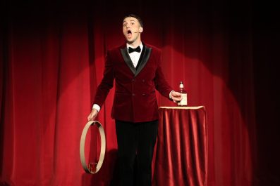 A man, Taylor Morgan, in a red velvet tuxedo on stage. He is carrying a hoop. Next to him on a pedestal is a bottle. The curtains behind him is also red velvet.