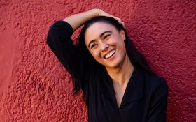 A young woman with black hair and wearing black has her head on an angle. She is smiling and has on hand on her head.