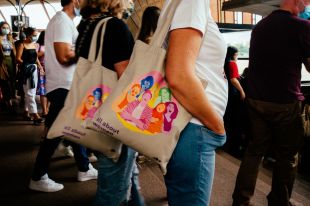 A crowd seen with two people carrying tote bags that say 'All about women' on them with a colourful illustration of a group of women.