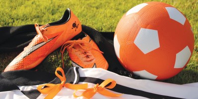 A pair of orange football shoes next to an orange and white football.