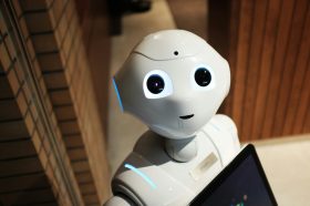 The head and shoulders of a white robot with black eyes seen from above. It's looking up at the camera. AI