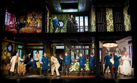 Interior cutout of a two-storey house in a production of 'The Barber of Seville'. There are people crowded in each room.