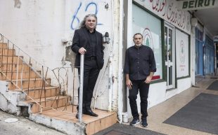 Artist Khaled Sabsabi (right, a middle-aged man wearing all black) and curator Michael Dagostino (left, a middle-aged man with long gray hair wearing all black) for the 2026 Venice Biennale. They are standing at the corner of a graffitied shopfront.