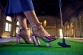 'Swingers – The Art of Mini Golf' has been announced as the first event for RISING 2025. The photo is a close-up of someone wearing glamorous high heeled shoes, standing on a fake putting green. Their right foot rests on a golf ball and they are leaning on a mini golf club. The dramatic space of the Flinders Street Station Ballroom is visible in the background.