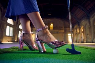 'Swingers – The Art of Mini Golf' has been announced as the first event for RISING 2025. The photo is a close-up of someone wearing glamorous high heeled shoes, standing on a fake putting green. Their right foot rests on a golf ball and they are leaning on a mini golf club. The dramatic space of the Flinders Street Station Ballroom is visible in the background.