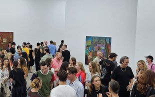 A large crowd gathers inside a gallery space with white walls, talking to each other.