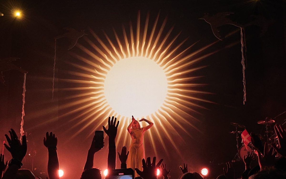 AURORA on stage in front of a large sun displayed on the screen behind her, while audiences hold up their hands as if reaching out.