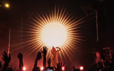 AURORA on stage in front of a large sun displayed on the screen behind her, while audiences hold up their hands as if reaching out.