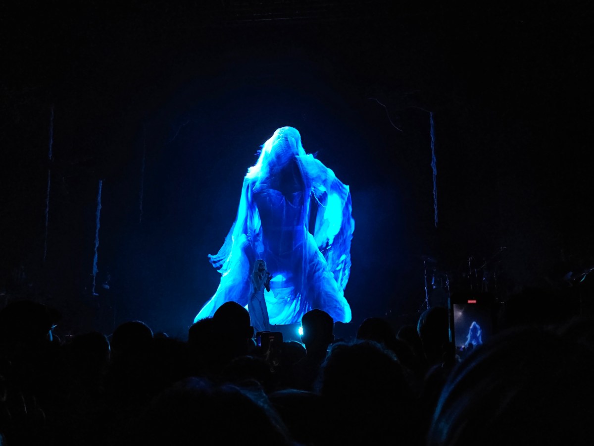 AURORA at Margaret Court Arena, Melbourne, 2025, featuring the singer on a dark stage with a blue wraith displayed on screen behind her.