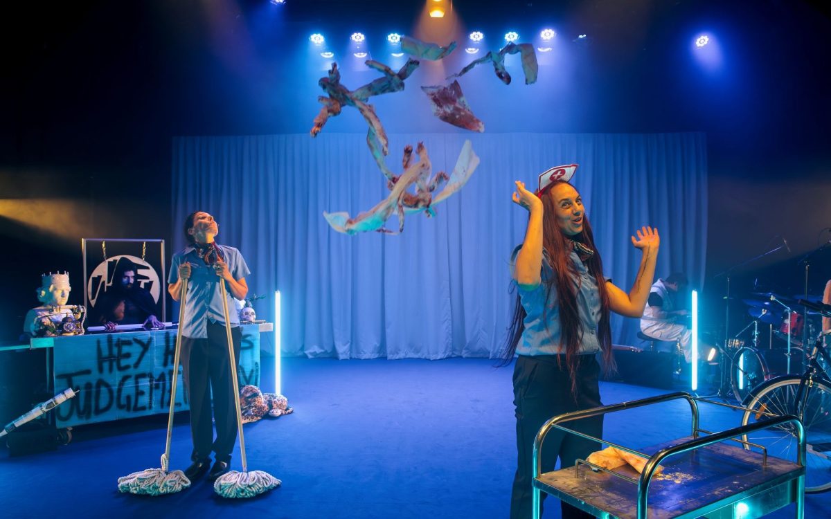 A scene from A Daylight Connection's 'A Nightime Travesty' at Malthouse Theatre for Asia TOPA 2025. Two Aboriginal women dressed as airline stewardesses stand on a blue-lit set as bloody rags fall from above. Behind them at stage right stands a sexy, shirtless, Aboriginal actor playing Death; at stage left, two musicians are visible.