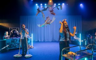 A scene from A Daylight Connection's 'A Nightime Travesty' at Malthouse Theatre for Asia TOPA 2025. Two Aboriginal women dressed as airline stewardesses stand on a blue-lit set as bloody rags fall from above. Behind them at stage right stands a sexy, shirtless, Aboriginal actor playing Death; at stage left, two musicians are visible.