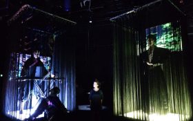 Opera for the Dead performance shot, featuring two movable stages, each with a performing standing at its centre, slightly obscured by black stingy curtains. The image is dark other than the stage being slightly illuminated from below.