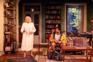 An elderly woman (Nancye Hayes) is standing in a living room filled with books. She's wearing a white dress. A 20 something man is seated on a couch (Shiv Palekar)