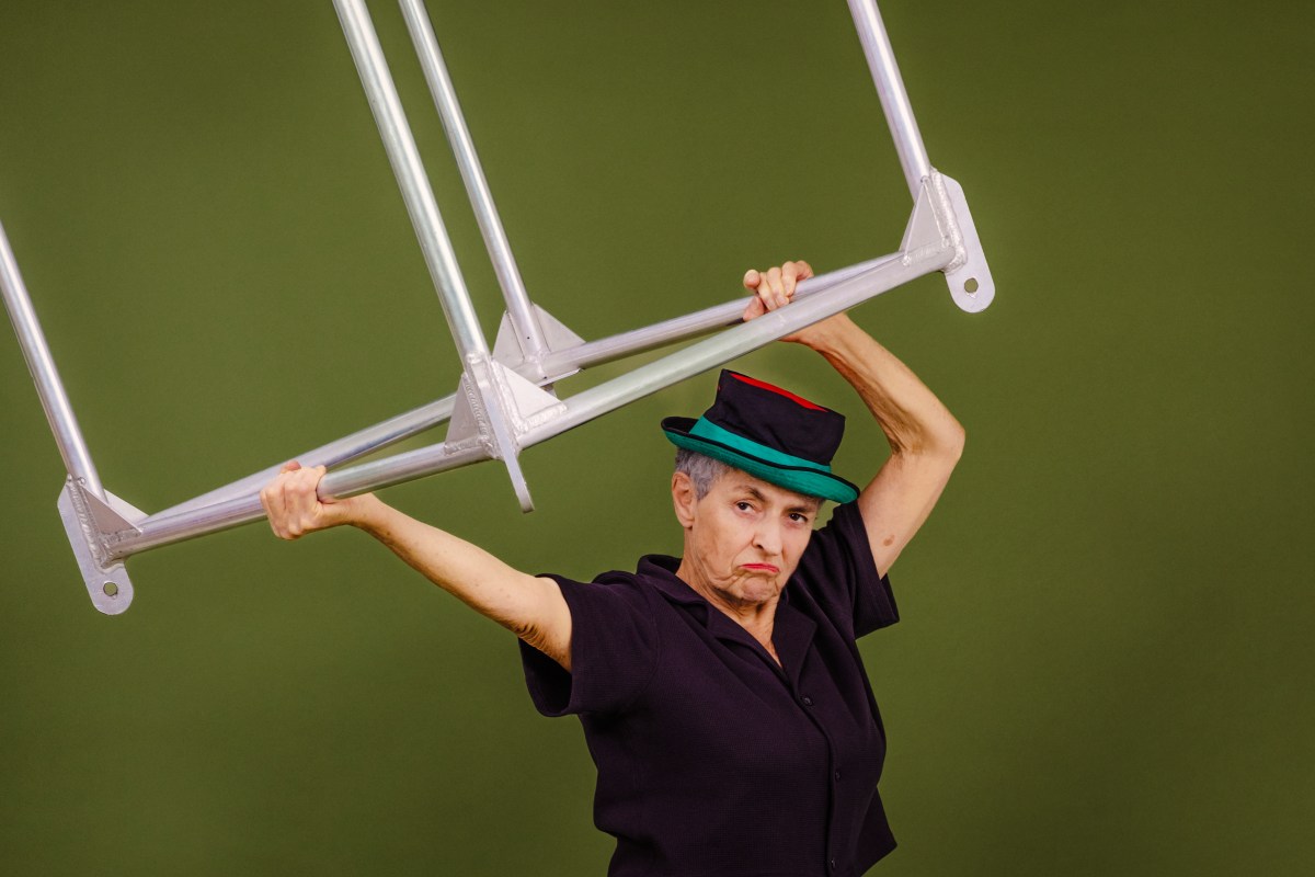 Against a green background a woman in a black shirt and short top hat holds up a metal frame and scowls at the camera. Small Acts of Resistance, Women's Circus.