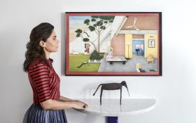 Photo of a woman with light brown skin and dark brown hair in side profile, standing to the left of a high countertop and a figurative painting of a suburban house on the wall.