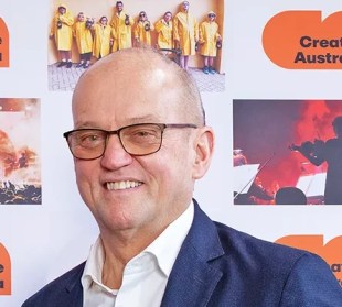 Creative Australia CEO Adrian Collette AM. A middle-aged, fair-skinned man with a shaved head, and wearing glasses and a blue suit, smiles at the camera.