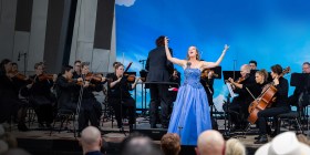 An opera diva in a big blue dress has her arms up in the air and a small orchestra behind her. Opera For All. Dimity Shepherd.