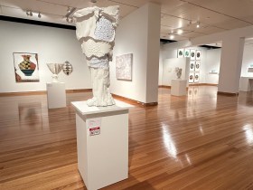 gallery view with large white ceramic pot on a stand and artworks in background. CMAG