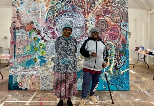 Two first nations women standing in front of a painting. Aboriginal art.