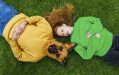 Lucy Heffernan's 'Dog People' as part of MERRIGONGX 2025. A photo of two figures lying on the grass, one is a woman with pale skin and red hair, wearing a green sweater, and another is a figure with a furry dog head and human body, wearing a yellow sweater.