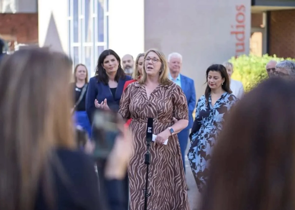 Woman in patterned dress surrounded by crowd giving a speech, outdoors.