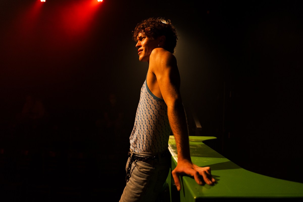 A young man in a singlet standing in profile and leaning his arms back on a green bench.