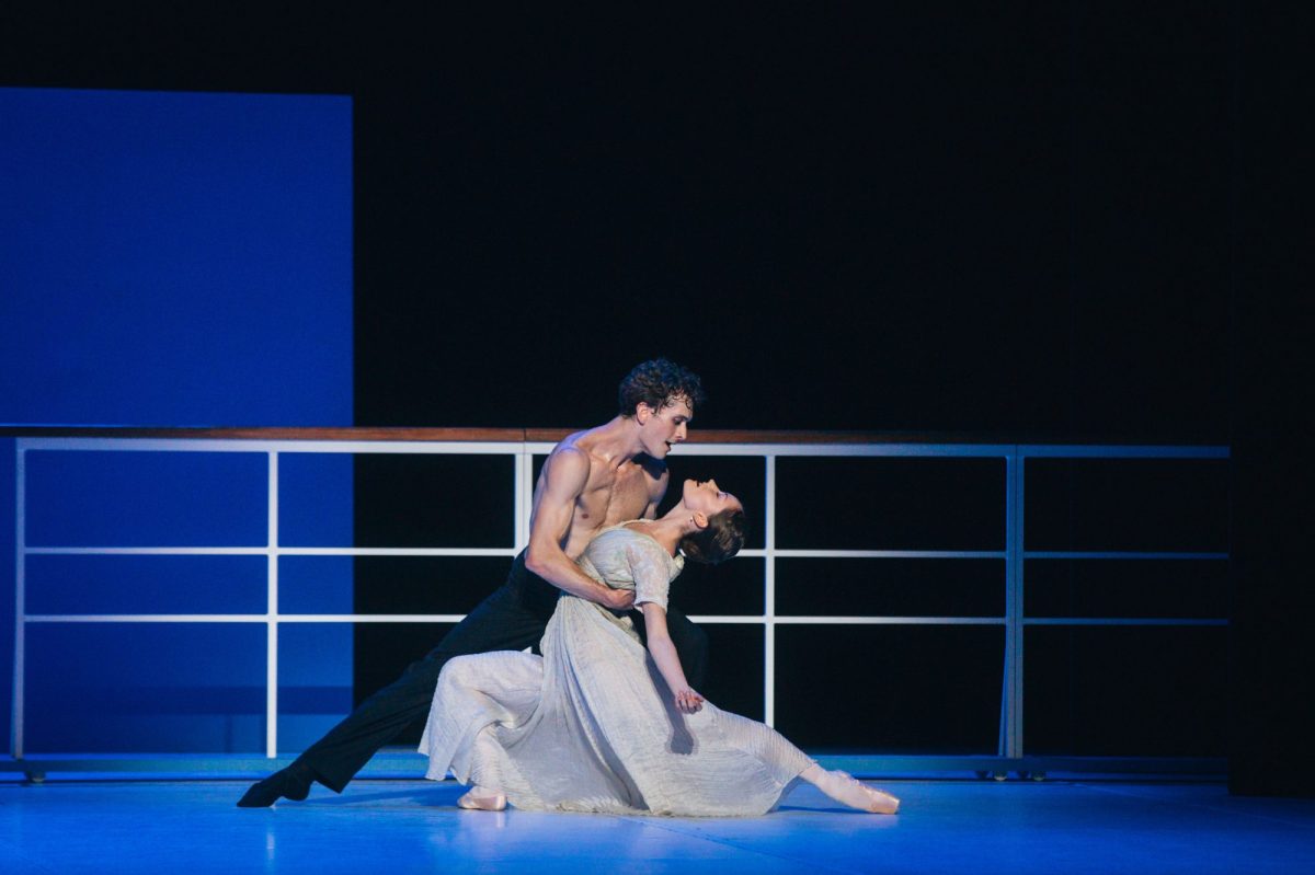 A bare-chested man with black pants is holding onto a woman in white. There is a white railing behind them.