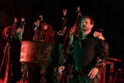 Hugh Sexton as Macbeth. He is dressed in various rags and furs. Behind him are the three witches around a cauldron.