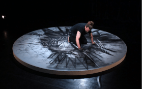 Thomas Buchanan is the inaugural Brown Falconer Artist in Residence program. A man kneeling on the ground on top of a round platform making expressive marks with black pigment.