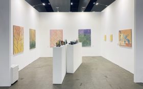 LON Gallery Booth C2 with a duo presentation of Jeremy Eaton and Stephen Benwell, installation view, Melbourne Art Fair 2025. A white walled space inside a grey convention centre. On the walls are canvases of soft yellow, green, orange and purple hues, while small earthenware sculptures are lined up on a plinth in the centre of the space.