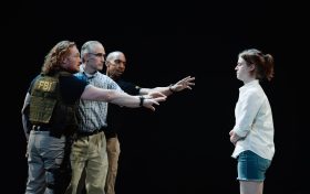 Two men, one with a FBI vest, are on the left, with their arms pointing to a woman on the right.