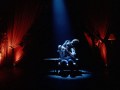 Two figures in shadowed darkness in the middle of the stage holding onto each other. Heavy drapery surround them with red tones.