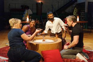 An African Australian woman with long dark haired tied back, a Caucasian woman with a messy blonde bun, an Indian man with a black bun and a Caucasian man with short blond hair sit around a pit with sand in it drinking espresso martinis on stage set decorated like a Turkish restaurant. Never Have I Ever, Melbourne Theatre Company.