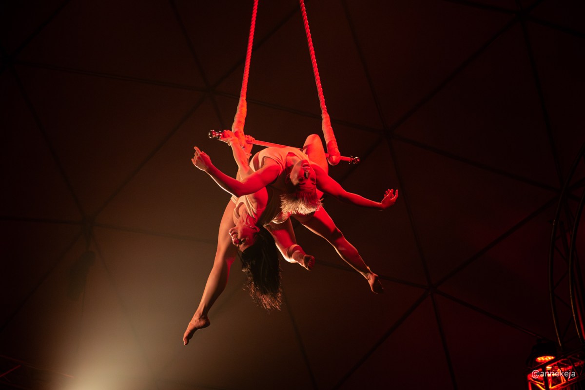 Two female performers are entwined on a trapeze.