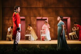 A man and woman facing each other. They are both dressed in Regency costumes. In the background are four other characters moving chairs around.