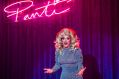 Rory O'Neill as Panti Bliss in THISISPOPBABY's 'If These Wigs Could Talk'. A 56-year-old drag queen in a blonde wig and grey dress gestures emphatically mid-speech. A neon sign of the word 'Panti' is visible behind her.