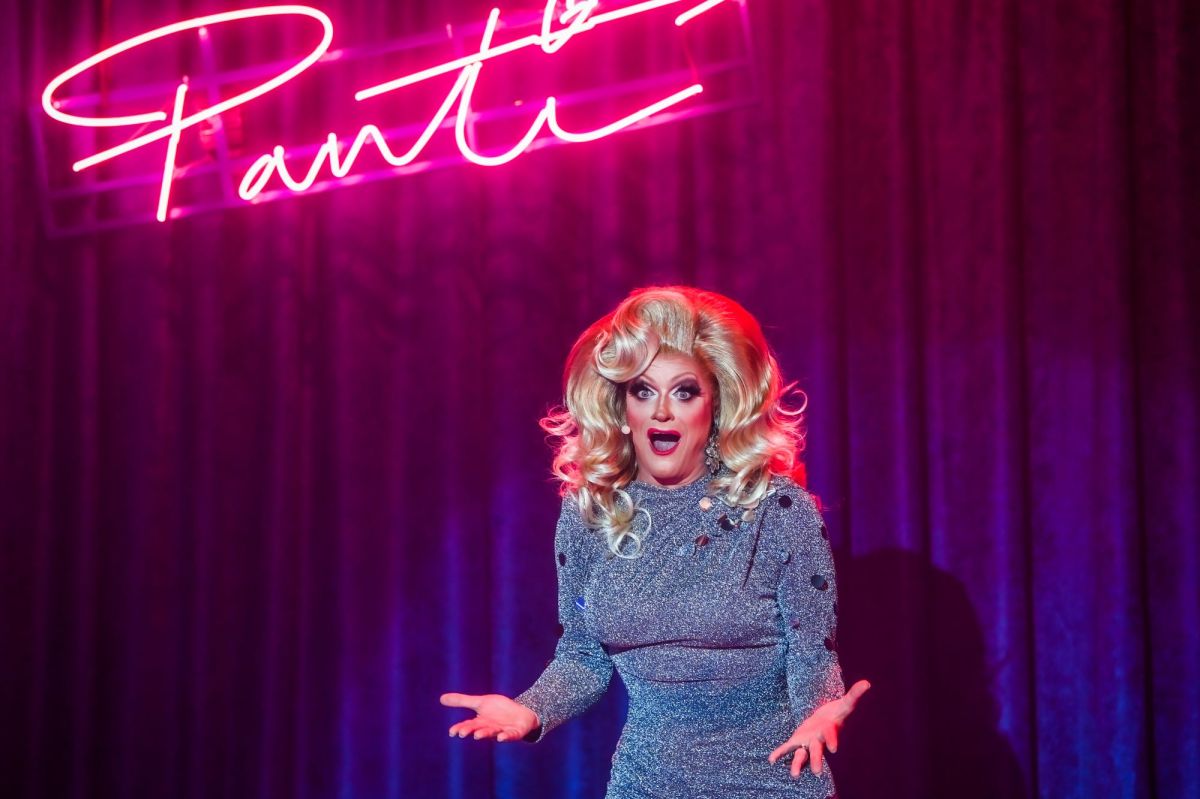 Rory O'Neill as Panti Bliss in THISISPOPBABY's 'If These Wigs Could Talk'. A 56-year-old drag queen in a blonde wig and grey dress gestures emphatically mid-speech. A neon sign of the word 'Panti' is visible behind her.