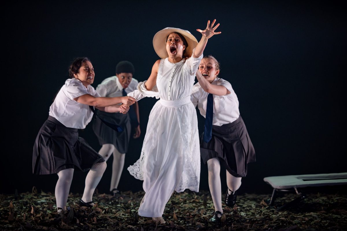 Three young girls in school uniform are holding back their teacher, who's dressed in white, with a straw hat.