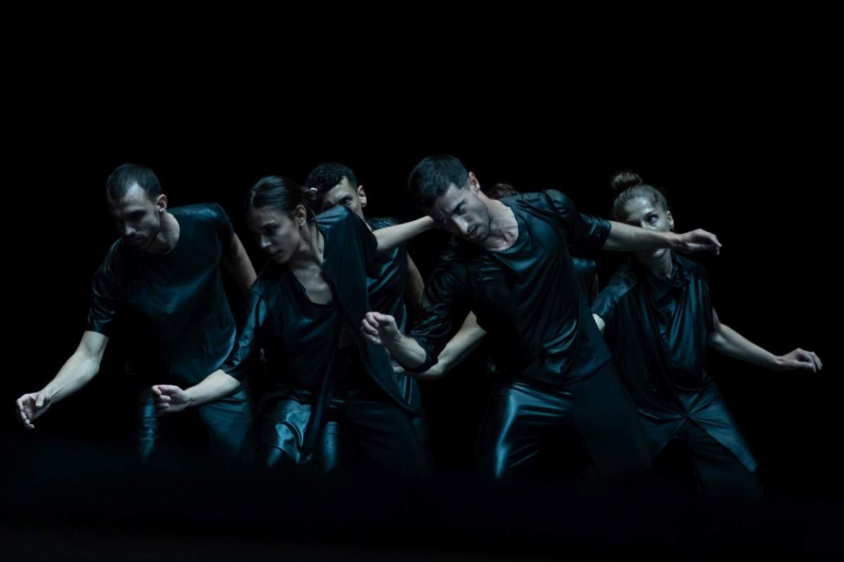 Larsen C: six dancers in metallic grey/black costumes pictured in synchronous movement bending sideways against a black backdrop.