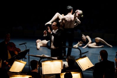 Conductor Umberto Clerici is conducting musicians in foreground. Behind him are several performers on the ground in different poses.