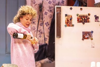 Natalie Bassingthwaighte in 'Shirley Valetine.' She has frizzy blonde hairstyle and is wearing a pink jumper. She is opening a champagne bottle next to a fridge.