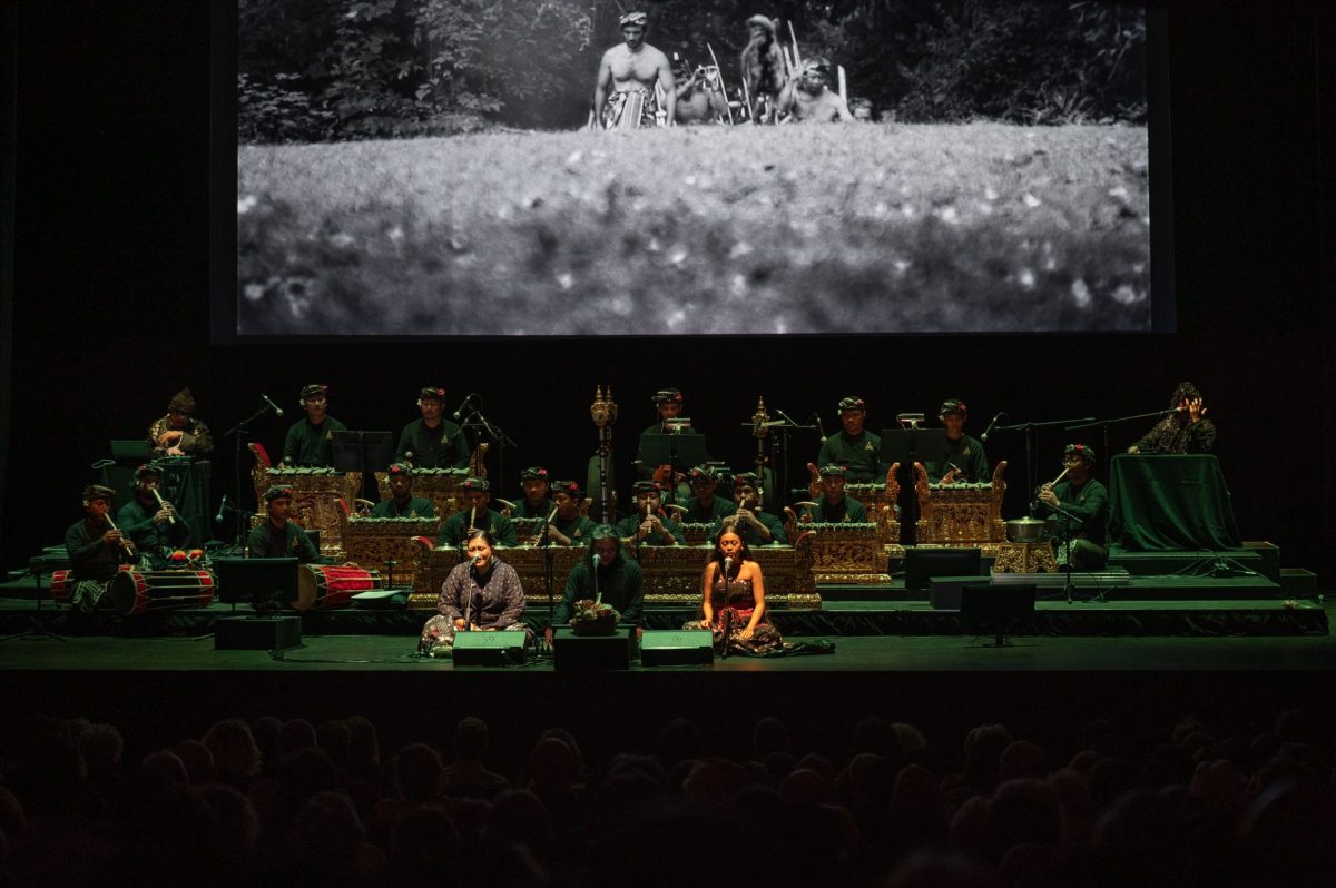 A large screen depicting a black and white screen showing a group of people in a field. There are musicians playing in the foreground.