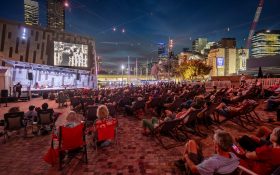 A crowd sitting on chairs under the night sky in federation square, an open-air area with a large screen.