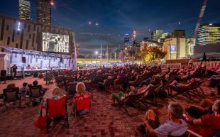 A crowd sitting on chairs under the night sky in federation square, an open-air area with a large screen.