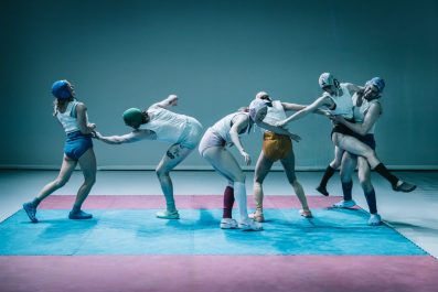 Six dancers wearing singlets and undies and skull-fitting caps are in various states of contortion.